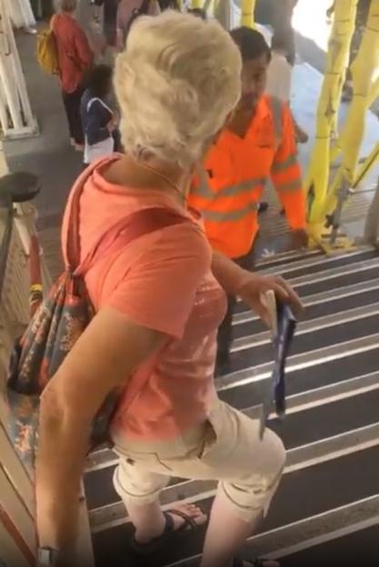 An elderly woman with blonde hair, wearing a pink shirt and white capris, attempts to climb stairs at a train station while a man in an orange reflective vest stands in front of her.