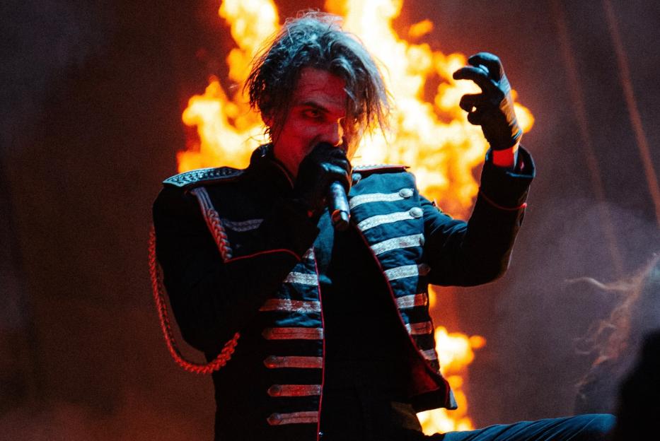 Gerard Way of My Chemical Romance performing in a military-style jacket with flames in the background.