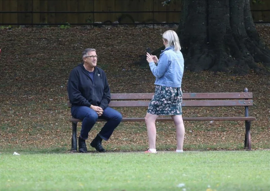 Graeme Hall sits on a park bench while Nicola Worth takes a video of him with her phone.