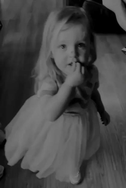 A young child with light hair wearing a fluffy dress, kneeling on a wooden floor, with one hand near their mouth.
