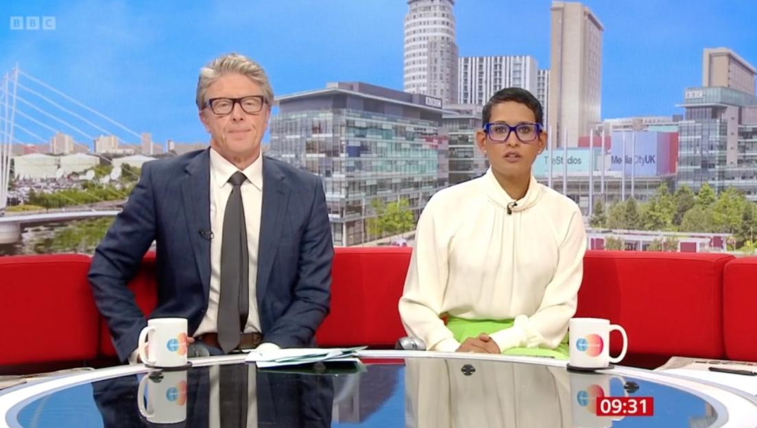 Two BBC anchors sitting at a desk with mugs, in front of a city skyline backdrop.