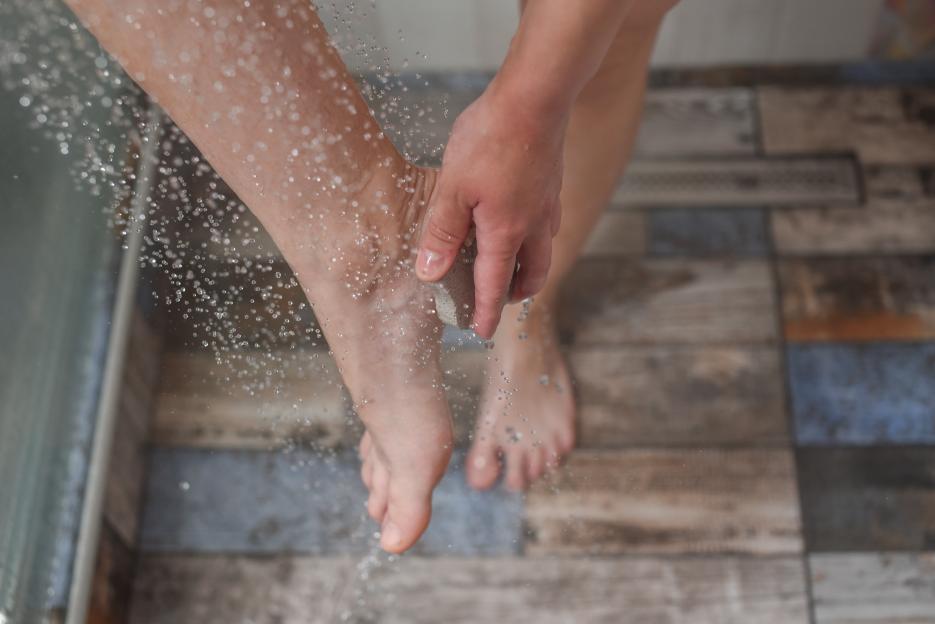 A person scrubbing their ankle with a pumice stone under shower water.