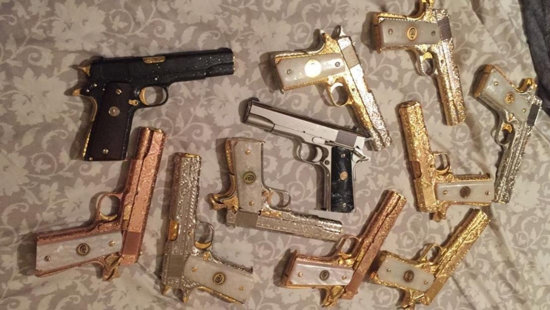 A collection of handguns, many of them highly customized with gold and silver plating and intricate engravings, laid out on a patterned fabric.