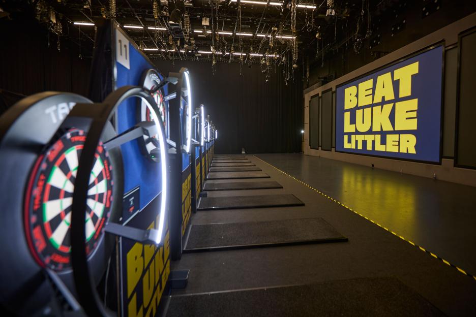 Darts boards illuminated on stage with a large "Beat Luke Littler" sign in the background.