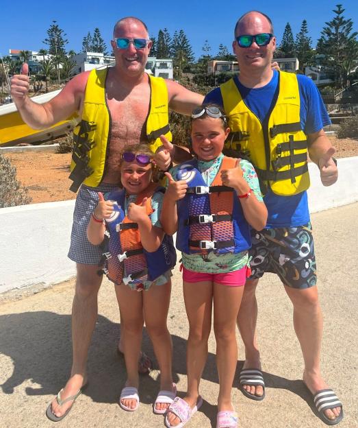 Two men and two girls in life vests give a thumbs-up.