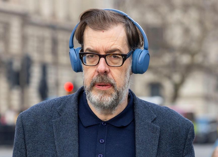 Richard Osman wearing glasses and blue headphones, looking forward.