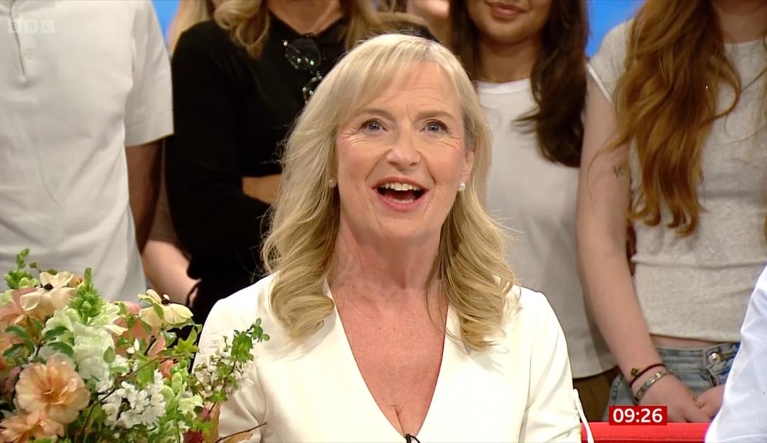 Carol Kirkwood smiling and looking up, with a bouquet of flowers next to her and people in the background.