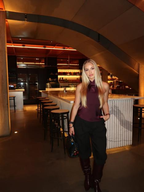 A woman with long blonde hair and a plum-colored top stands in a bar.