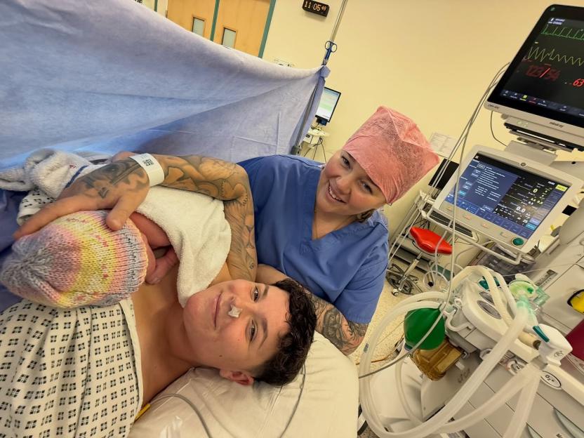 A person with tattoos holding a newborn baby wrapped in a blanket and wearing a colorful knitted hat, next to a smiling medical professional in a blue scrub top and pink scrub cap.