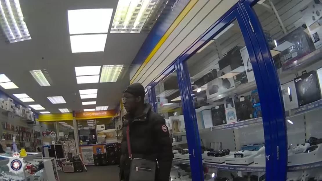 A man in a black jacket and beanie looks around an electronics store, with video game consoles and accessories on display in glass cases.