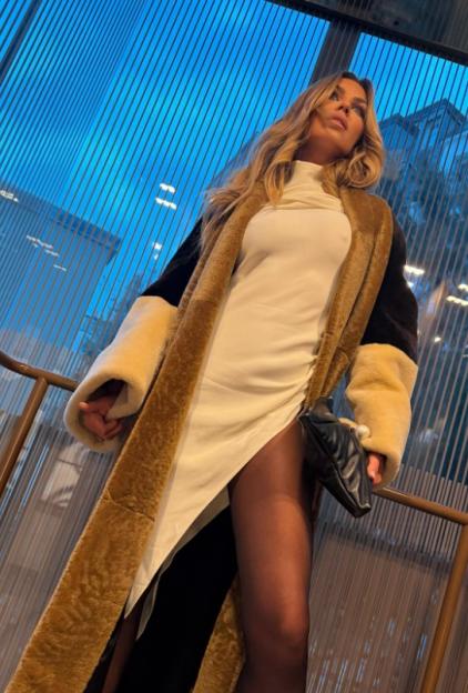 Abbey Clancy standing with a long gold and cream coat over a white dress with a high slit, holding a black handbag.