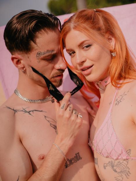 Couple with tattoos, the man holding sunglasses while the woman wears a pink bikini top.