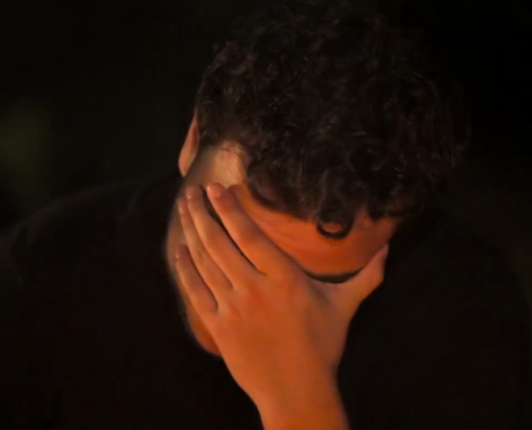 A man with curly hair covering his face with his hand in a dark setting.