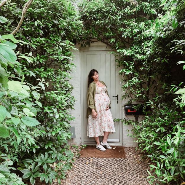A pregnant woman in a white floral dress and green jacket stands in a leafy doorway.