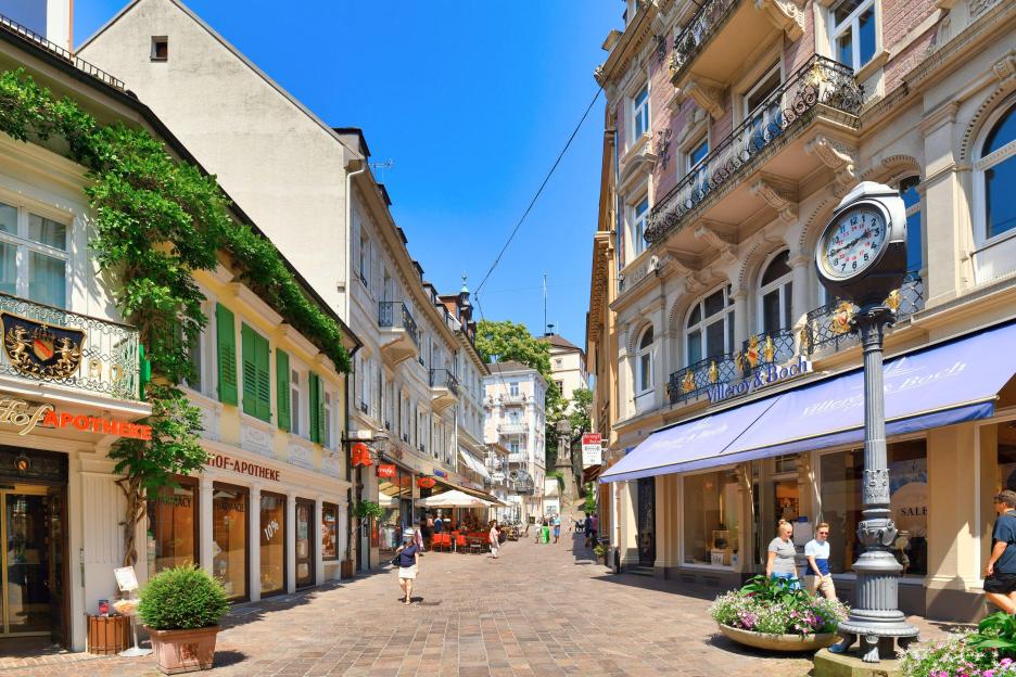 A narrow pedestrian street with shops, balconies, and a decorative street clock.