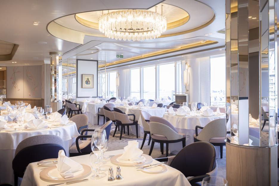 Interior of the Princess Grill restaurant on board the Queen Anne cruise ship, featuring set tables, plush chairs, and large windows.