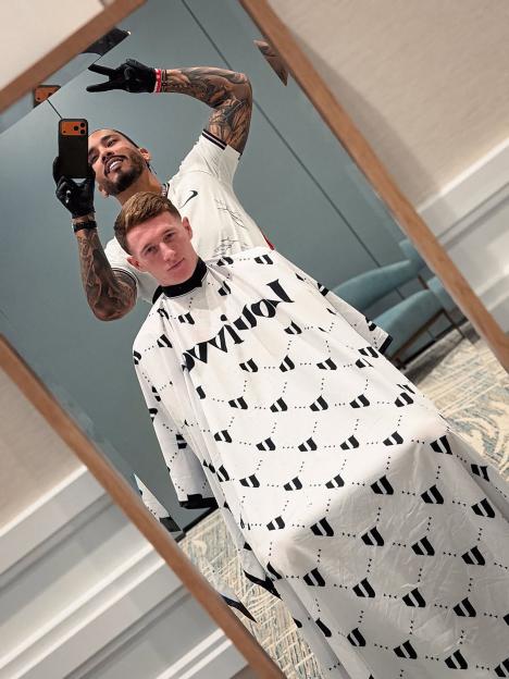 A mirror selfie showing a tattooed barber in a white Nike shirt giving the "peace" sign, while a person in a white haircut cape looks into the mirror.
