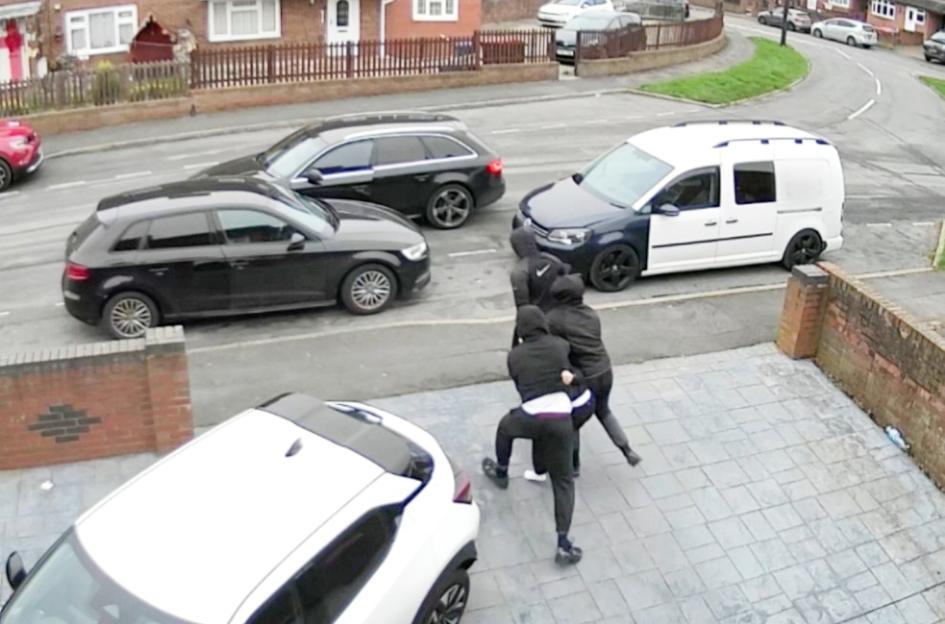 Two men in dark clothing grappling on a paved driveway, with cars on the road behind them.