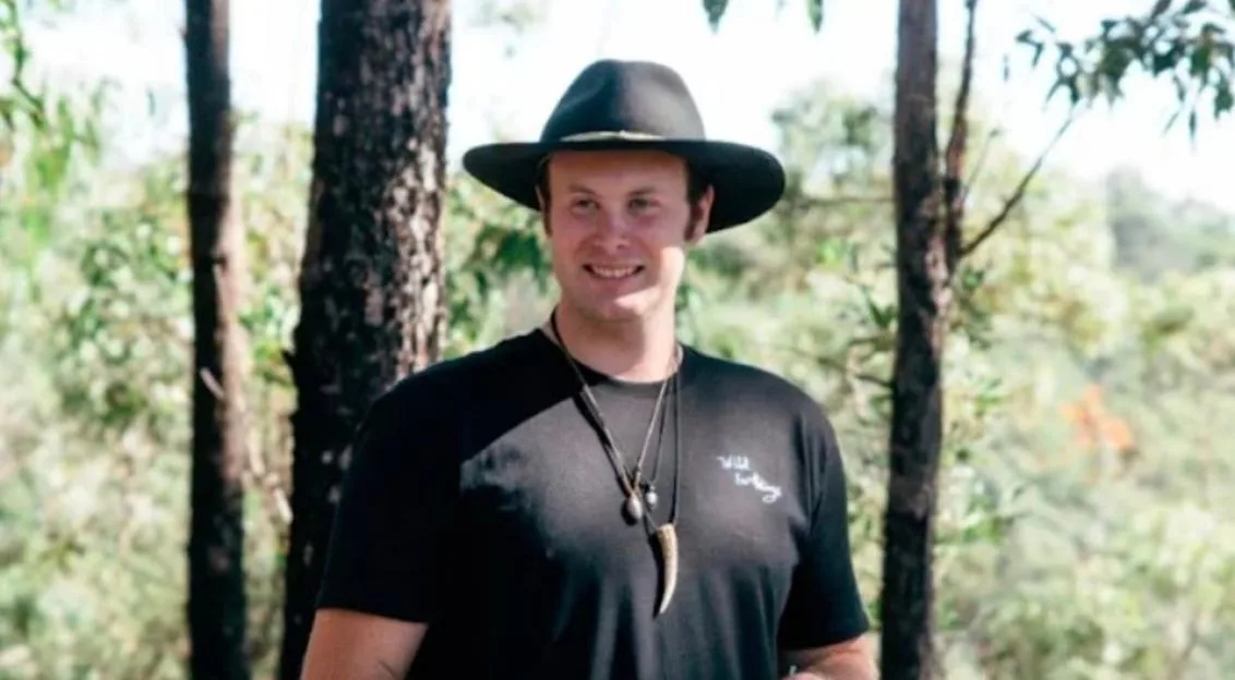 A man in a black hat and black t-shirt with a necklace with a large horn-shaped pendant smiles slightly in a forest.