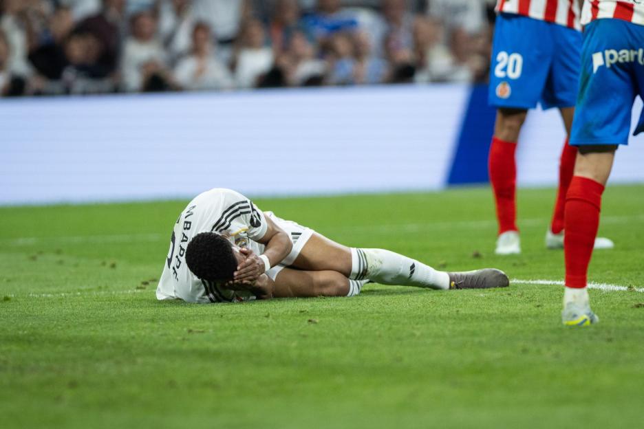 A Real Madrid CF player in a white jersey with "MBAPPE" on the back lies on the ground, holding their head in their hands, while two Girona players in red and blue stand nearby.