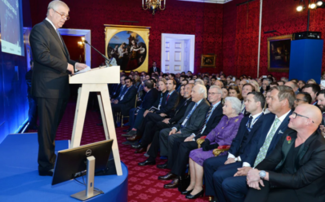 Prince Andrew speaking at a podium to an audience seated in a red room.