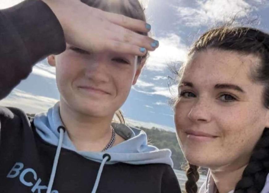 A young girl with blue nail polish shielding her eyes from the sun next to a woman with braided hair.