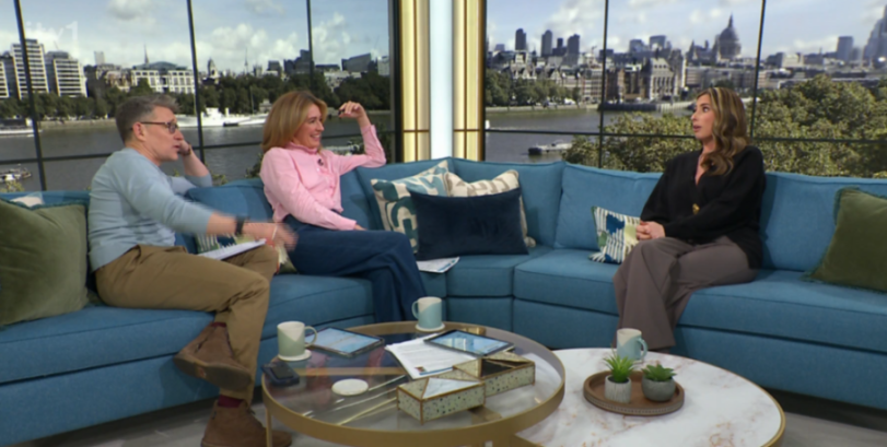 Three people on a blue sofa in a talk show setting with a city skyline backdrop.