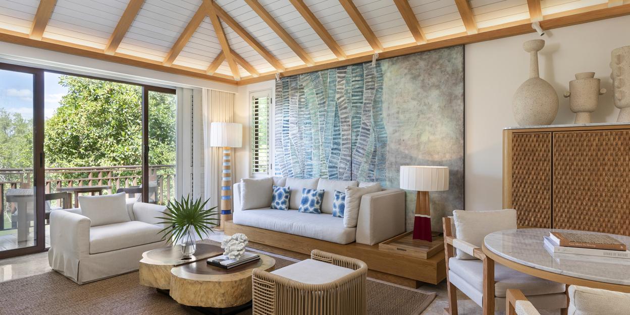 A living room with a white couch and chairs, wood-beam ceiling, and large sliding doors opening to a lush green outdoor area.