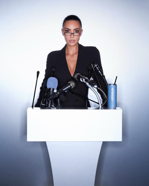 Kim Kardashian in a black suit with a plunging neckline and glasses, standing behind a podium with microphones and a blue "UPDATE" drink can.