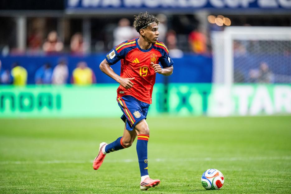 A player in a red and blue uniform with the number 19 on his back runs next to a soccer ball on a green field.