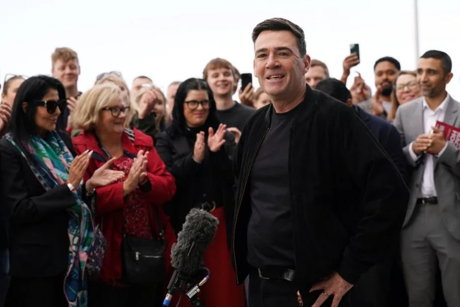 Andy Burnham, dressed in a black t-shirt and jacket, stands smiling in front of a crowd of people who are clapping.