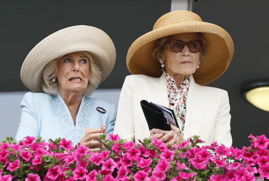 Queen Camilla and another woman in large hats watching horses from a balcony.