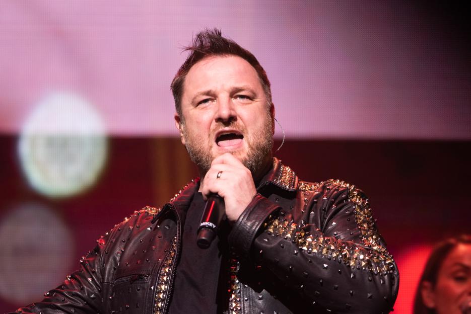 Mikey Graham of Boyzone singing into a microphone on stage, wearing a black studded leather jacket.