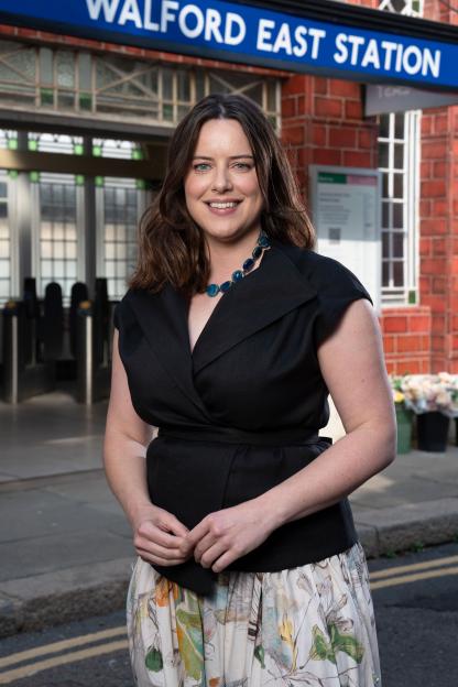 Michelle Ryan as Zoe Slater in front of a "Walford East Station" sign.