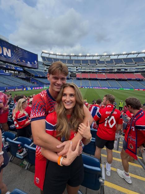 A man and woman wearing red shirts with the Norwegian flag embracing each other at a stadium during what appears to be a FIFA World Cup event.