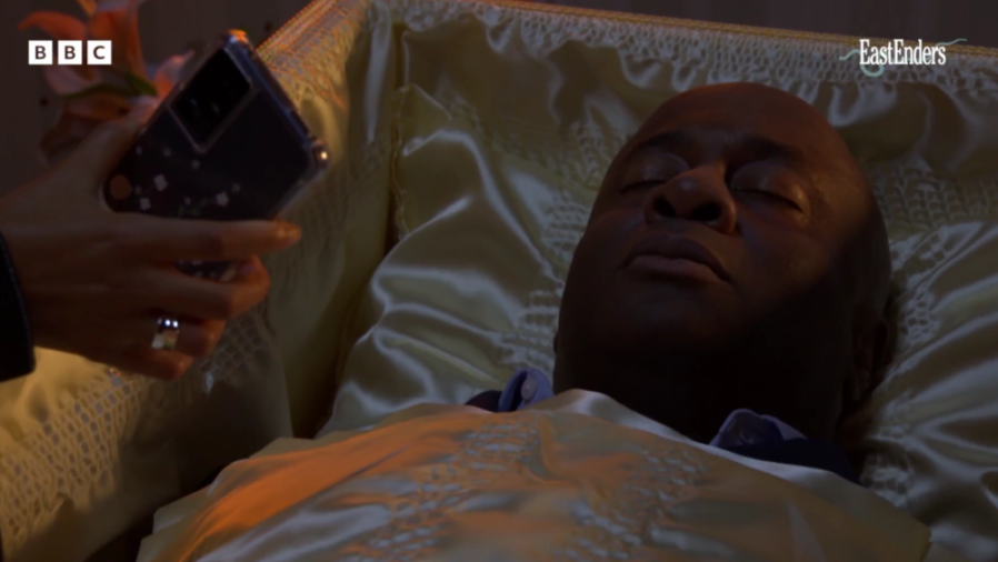 A hand holding a smartphone to photograph a man lying in a satin-lined casket.