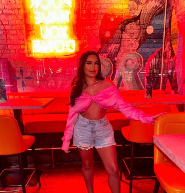 Mia Pritchard wearing a pink top and denim shorts in a bar lit with red neon lights.