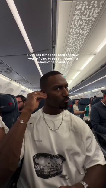 A man on an airplane in a white t-shirt and silver chain looking at the camera with the text overlay "POV: You flirted too hard and now you're flying to see someone in a whole other country".