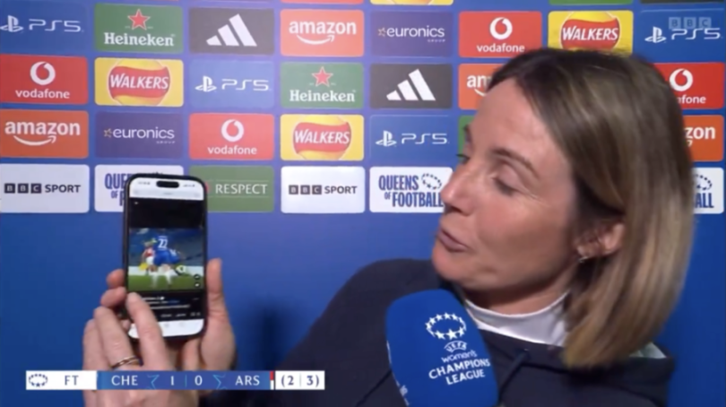 A woman holds up a phone displaying a soccer game replay during a post-match interview.