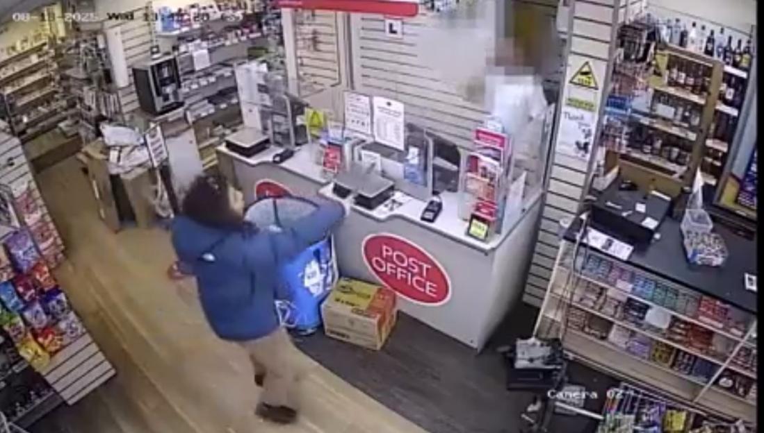 A person in a blue jacket reaching into a blue bin at a Post Office counter, with shelves of items in the background.