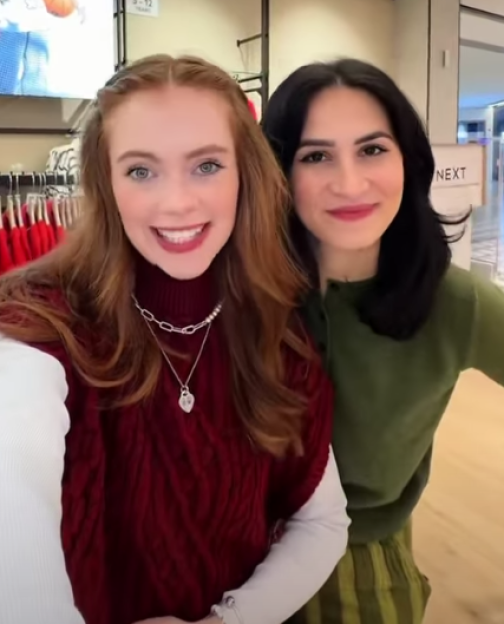 Two women smiling in a clothing store, one with red hair and a red top, the other with dark hair and a green top.