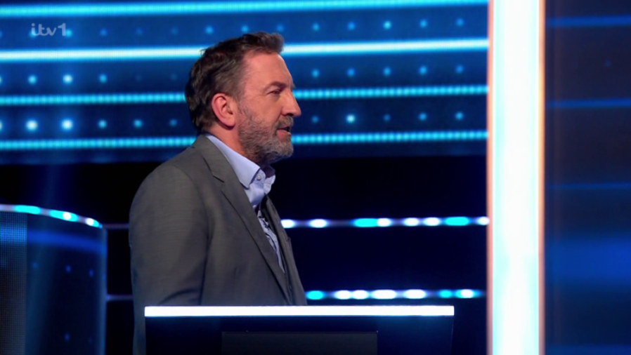 A man in a suit and collared shirt stands to the left of the frame, facing right, against a blue, illuminated background.