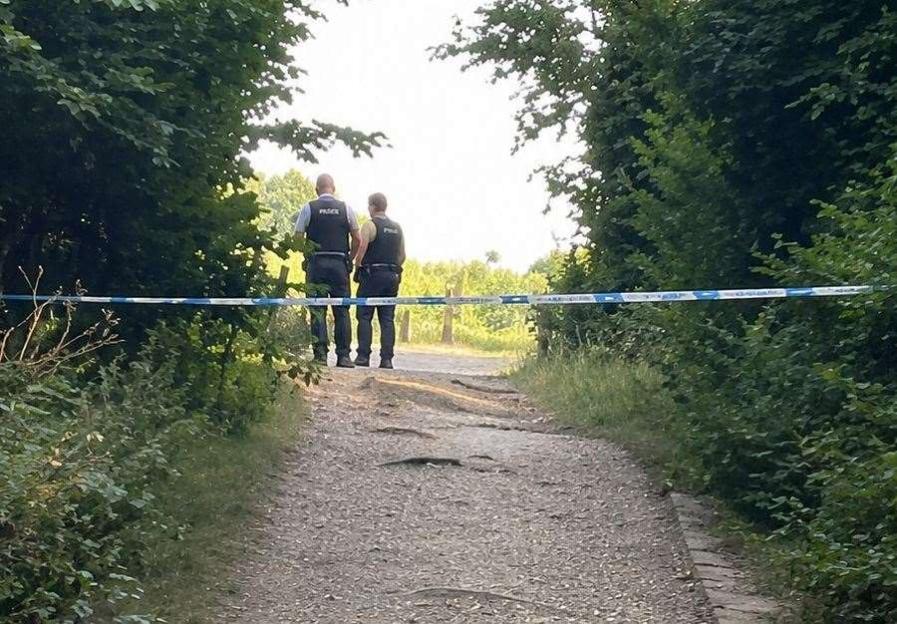 Two police officers stand on a dirt path in a wooded area, behind blue and white police tape.