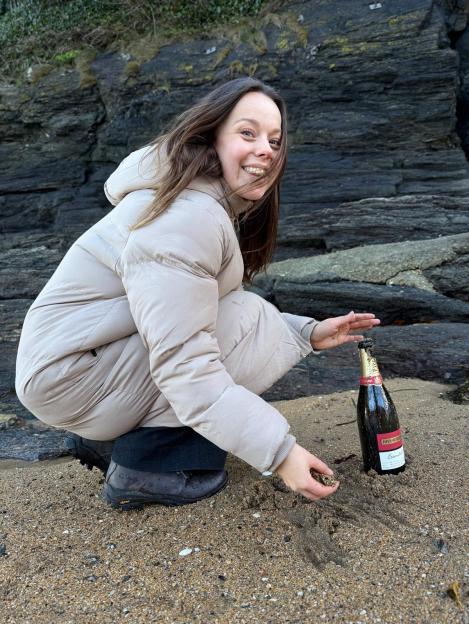 Evie Pickerill celebrating her engagement on a beach, posing with an open bottle of champagne.