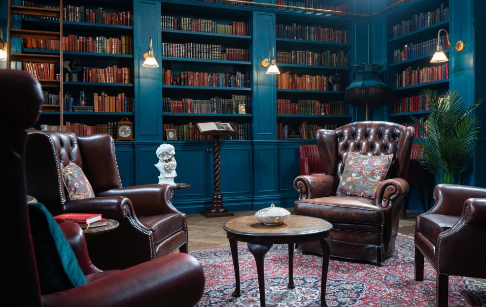 A blue-paneled library with brown leather armchairs, a wooden side table, and a decorative rug.