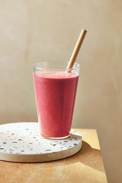 A pink smoothie with a straw in a clear glass on a round stone coaster.