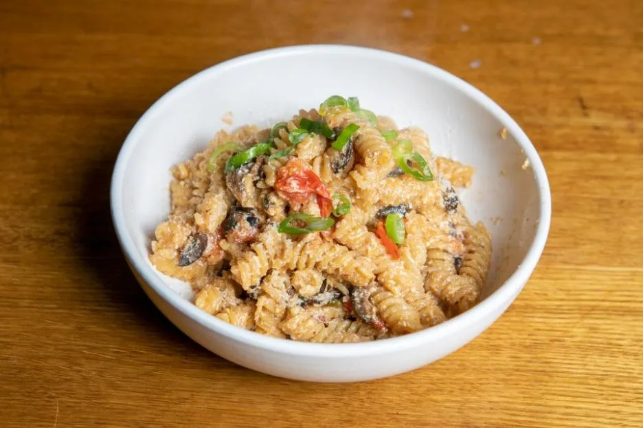 A white bowl of fusilli pasta with tomatoes and olives on a wooden table.