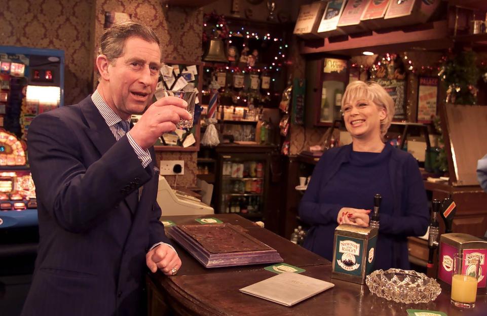 Prince Charles holding a drink with actress Denise Welch behind the bar of the Rovers Return pub set.