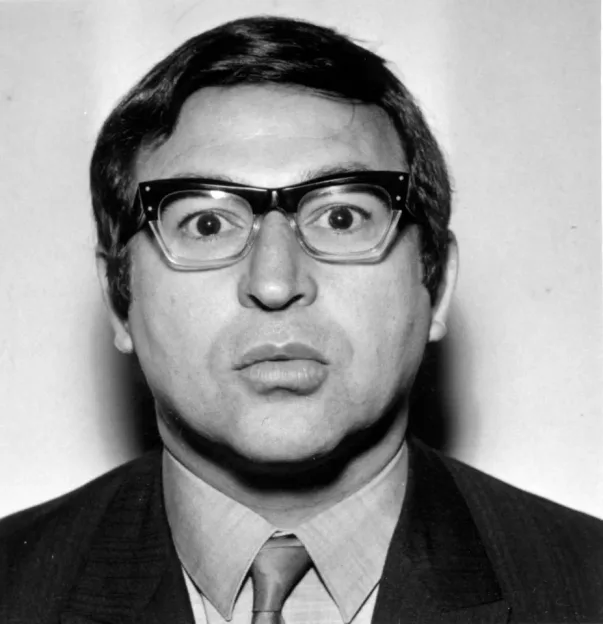 Black and white portrait of a man wearing thick-rimmed glasses and a suit, making a funny facial expression.