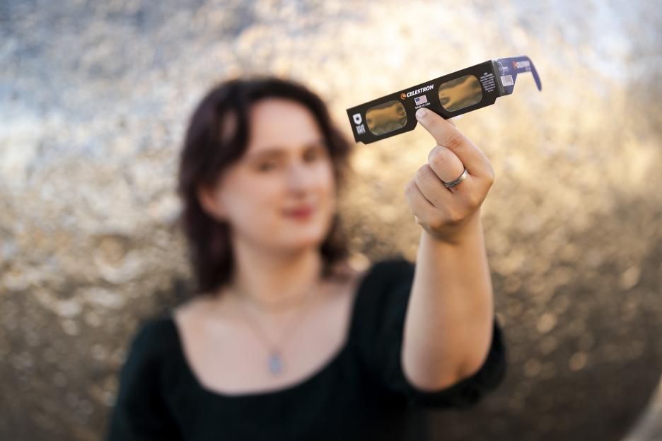 A young woman holds up Celestron eclipse glasses, which are in focus, while she is out of focus in the background.